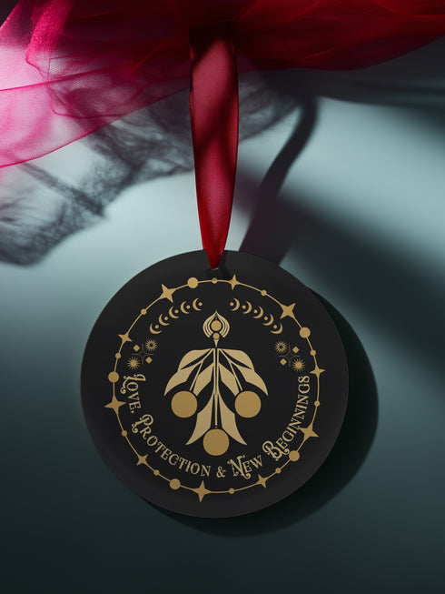 Round metal ornament featuring golden mistletoe design with Art Deco border incorporating stars and moon phases. 3.5-inch diameter piece with "Love, Protection & New Beginnings" text and crimson ribbon. Double-sided design on black background.